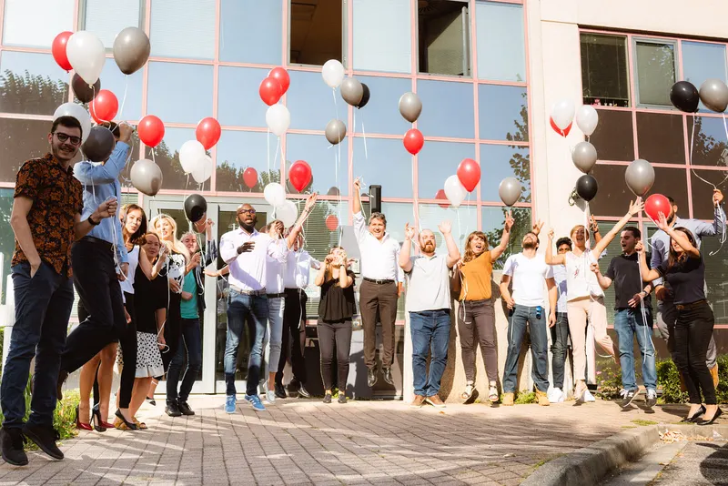 Équipe réunie devant ses locaux, ballons rouges, noirs et blancs, bras levés et sourires