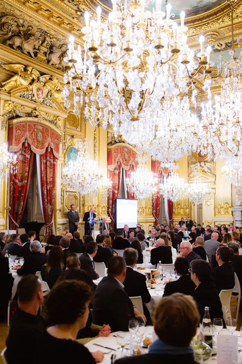Dîner de gala dans un salon doré aux lustres de cristal, convives attablés face à un intervenant