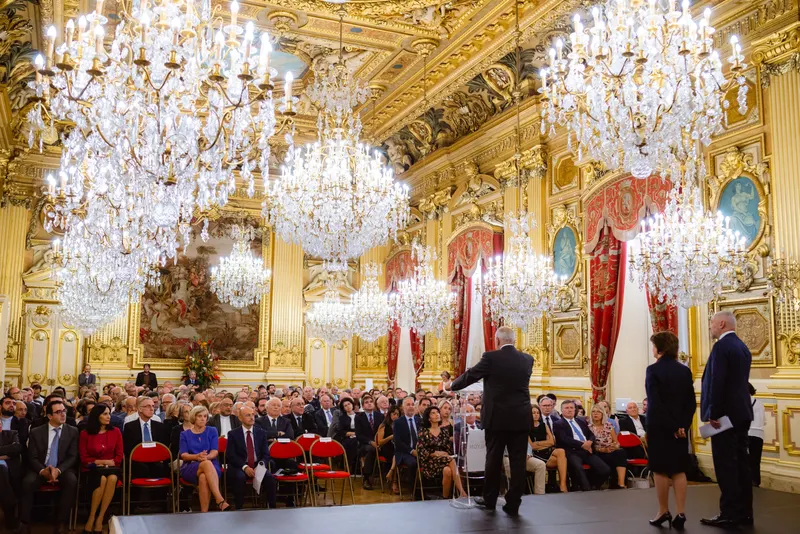 Conférence dans un salon historique, intervenant devant un public sous des lustres de cristal