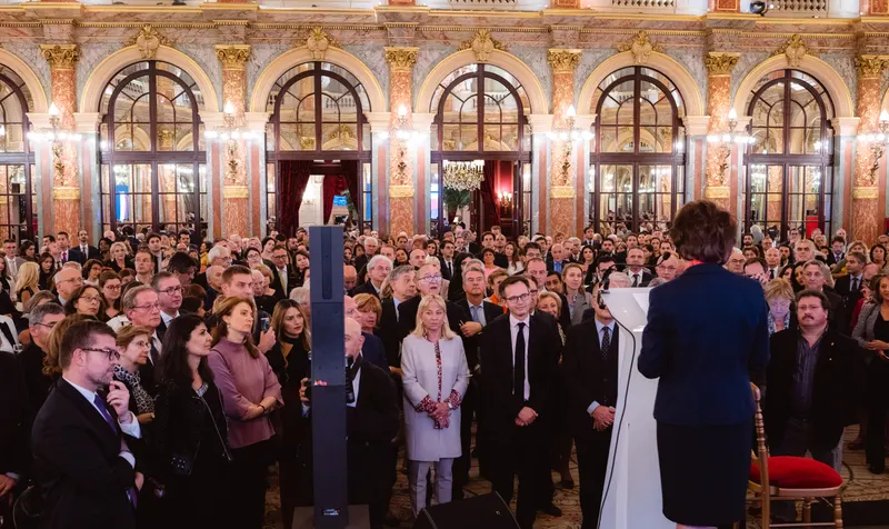 Public nombreux écoutant une oratrice au pupitre dans une salle ornée