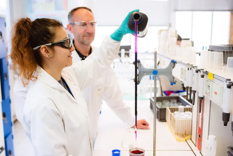 Technicienne de laboratoire versant un liquide avec une burette, un collègue derrière elle