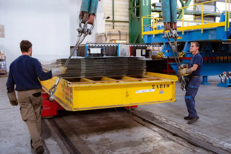Deux ouvriers guidant une charge de tôles suspendue à un pont roulant