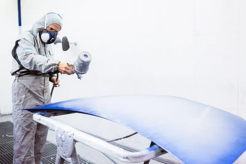 Peintre en combinaison grise pulvérisant de la peinture bleue sur une pièce de carrosserie
