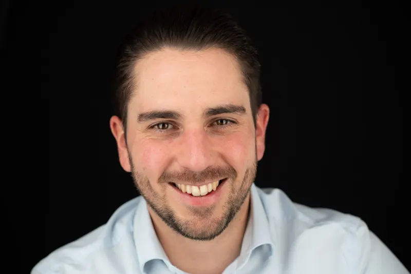 Portrait d’un homme barbu au grand sourire, chemise bleu clair, fond noir